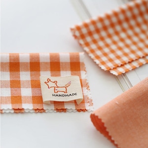 Puede incluir: Tela de cuadros naranja y blanca con una pequeña etiqueta blanca que dice "HANDMADE" y tiene un zorro de dibujos animados con un corazón.