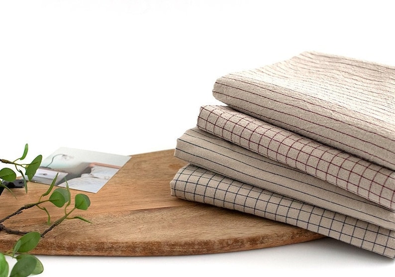 May include: Four beige and brown checkered fabric napkins stacked on a wooden cutting board. The napkins have different sized squares and are made of a soft, textured material.