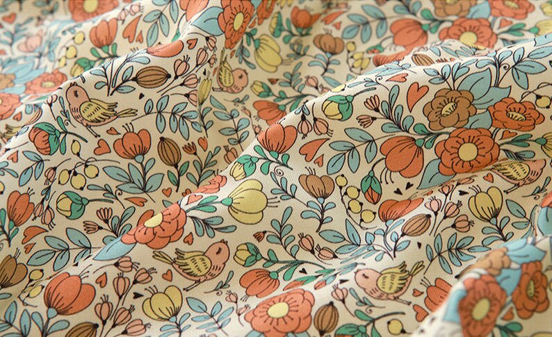 May include: A cream-colored fabric with a repeating pattern of orange and yellow flowers, green leaves, and small birds. The pattern is whimsical and charming.