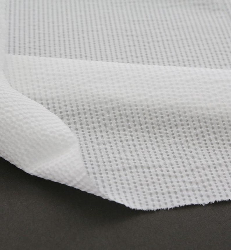 May include: White waffle-weave fabric with a fine, textured pattern. The fabric is folded and draped over a dark surface.