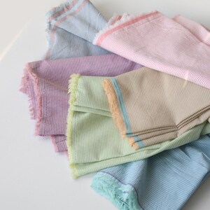 May include: Five striped fabric swatches in different colors, including light blue, pink, purple, green, and beige. The fabric has a frayed edge.