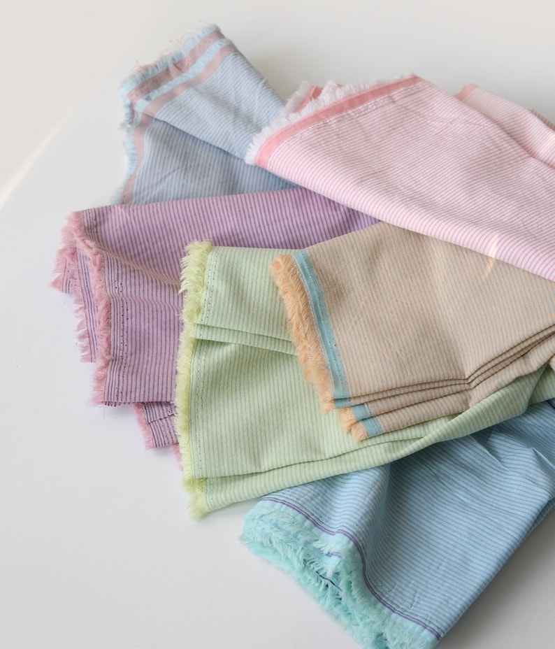 May include: Five striped fabric swatches in different colors, including light blue, pink, purple, green, and beige. The fabric has a frayed edge.