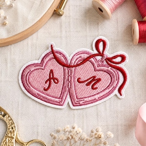 May include: An embroidered patch shaped like two connected pink hearts with a red ribbon bow. The left heart features the letter A and the right heart features the letter M in red thread. The patch has a white border and is placed on fabric.