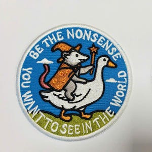 May include: Two circular embroidered patches featuring a gray opossum wearing an orange wizard hat and cloak, riding a white goose. The background is blue with a green ground and the text "BE THE NONSENSE YOU WANT TO SEE IN THE WORLD" in white.