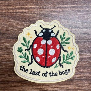 May include: An embroidered patch with a cream-colored background featuring a red ladybug with white spots, green leafy sprigs, and small yellow stars. The text "the last of the bugs" is stitched in black lettering along the bottom edge.