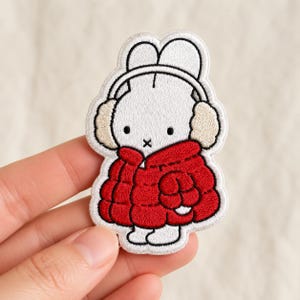 May include: An embroidered patch featuring a white rabbit character wearing beige earmuffs and a bright red puffer jacket. The design has a white border and black embroidered details for the eyes and mouth, creating a clear, graphic illustration.