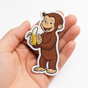 May include: An embroidered patch featuring a brown monkey character holding a yellow banana. The patch has a white stitched border and depicts the character with a smiling expression, light brown face, ears, hands, and feet, and dark brown fur.