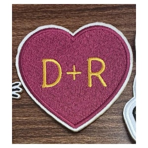 May include: A heart-shaped embroidered patch featuring a deep magenta fabric base with a white stitched border. The center of the patch displays the gold-colored embroidered letters D and R separated by a gold-colored plus sign symbol.