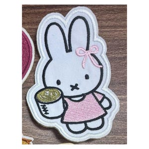 May include: An embroidered fabric patch featuring a white rabbit character with long ears, a pink bow, and a pink dress. The rabbit is holding a white mug with a green beverage inside. The design is outlined in black thread on a white background.