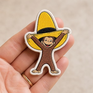 May include: An embroidered patch featuring a brown monkey character wearing a large yellow hat with a black band. The monkey is smiling and holding the brim of the hat with both hands. The patch has a white border and is shaped to the outline of the figure.