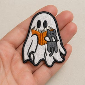 May include: An embroidered patch featuring a white ghost with black oval eyes. The ghost is holding an open orange book and a small gray cat. The entire design is outlined in a thick black border, creating a clear and defined shape for the patch.