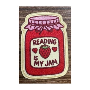 May include: An embroidered patch shaped like a jar of red strawberry jam with a light pink lid cover. The label features a central red strawberry illustration, two small red hearts, and the text Reading Is My Jam in dark brown lettering.