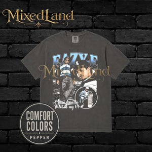 Puede incluir: Una camiseta de manga corta gris carbón con un estampado gráfico de Eazy-E. El diseño incluye el texto EAZY-E en azul, un retrato del artista y la frase Cruisin down the street in my 64. Se observa una etiqueta circular de Comfort Colors Pepper.