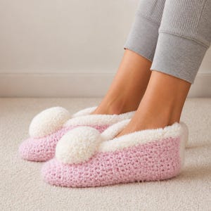 Puede incluir: Un par de zapatillas de punto rosa claro con un patrón de ganchillo texturizado y un pompón blanco grande y esponjoso en la parte superior de cada pie. Las zapatillas tienen un borde blanco suave alrededor de la abertura y punta redondeada.