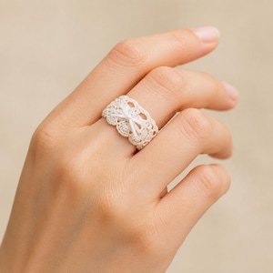 Puede incluir: Un anillo ancho de ganchillo en color blanco roto que presenta un intrincado patrón de encaje con bordes festoneados. Una cinta fina a juego está tejida a través del centro del ganchillo, atada en un pequeño y delicado lazo al frente.
