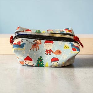 May include: A light blue zippered pouch featuring a repeating Christmas pattern of Santa Claus, reindeer, snowmen, pine trees, and wrapped gifts. The bag has a black zipper tape with a metallic gold zipper and a red pull tab on the side.