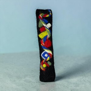 Puede incluir: Un adorno rectangular alto de lana fieltrada negra que presenta un diseño vertical en espiral de hilo multicolor en tonos rojo, azul, amarillo, verde y blanco, con pequeñas formas de fieltro integradas en la superficie.