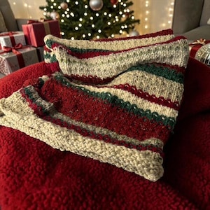 May include: A knitted cowl scarf featuring horizontal stripes in cream, deep red, and dark green. The texture is chunky and consistent throughout the piece, showcasing a repeating pattern of varied knit stitches that create a soft, folded appearance.