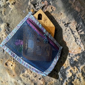 May include: A rectangular pouch with a dark blue mesh front and a purple zipper featuring a paw print charm. The pouch has a light-colored iridescent trim and contains a wooden house-shaped object with a small cutout and etched symbol.