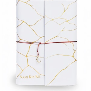 May include: A white rectangular notebook featuring a gold-colored crackle pattern across the cover. The notebook is secured with a thin dark red cord that includes a small gold-colored crescent moon charm. The text NAMI KIN KO is printed in gold.