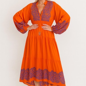 Puede incluir: Un vestido midi de color naranja vibrante con un escote en V profundo y mangas largas y voluminosas. La prenda está adornada con intrincados bordados morados a lo largo del escote, los puños y el dobladillo. Tiene un diseño de falda escalonada.