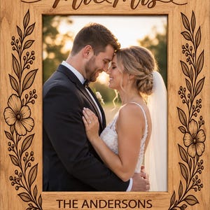 Pode incluir: Uma moldura retangular de madeira com vinhas florais gravadas a laser nas laterais, o texto "Mr & Mrs" no topo e "THE ANDERSONS" com a data "JUNE 16, 2021" na parte inferior, contendo uma fotografia de um casal a abraçar-se.