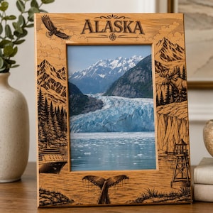 Könnte beinhalten: Ein heller Holzbilderrahmen mit lasergravierten Illustrationen alaskischer Landschaften, darunter ein Adler, Berge, ein Elch, ein Kreuzfahrtschiff, ein Leuchtturm und eine Walflosse. Das Wort ALASKA ist oben mittig auf dem Rahmen eingraviert.