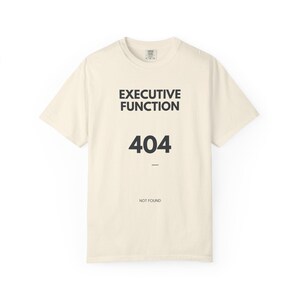 May include: A cream-coloured short-sleeve t-shirt featuring black text that reads EXECUTIVE FUNCTION, followed by the number 404, and the text NOT FOUND at the bottom. The shirt has a crew neckline and a standard fit with a simple graphic design.