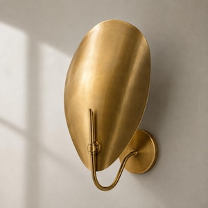 May include: A wall-mounted sconce featuring a large, oval-shaped brass shade with a brushed metallic finish. The light fixture includes a curved arm that connects the shade to a circular wall plate, creating a sleek and functional lighting design.