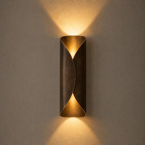 Pode incluir: Um candeeiro de parede cilíndrico com um acabamento em bronze escuro envelhecido. A luminária apresenta um design curvo sobreposto que emite uma luz dourada quente pelas aberturas superior e inferior, projetando feixes simétricos na parede.