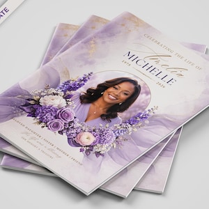 May include: A stack of funeral program booklets featuring a soft purple watercolor background with gold accents. The cover displays a circular photo of a smiling person, a floral arrangement of purple and white flowers, and the text CELEBRATING THE LIFE OF MICHELLE 1975-2026.