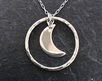 May include: A silver necklace with a hammered circle pendant and a crescent moon charm. The necklace is on a dark gray background.