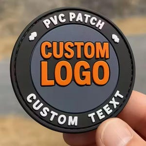 May include: A circular black PVC patch featuring a dark gray center with the orange text CUSTOM LOGO. The black border includes the white text PVC PATCH at the top and CUSTOM TEEXT at the bottom, separated by two small white cloud-like icons.