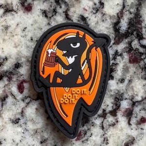 Könnte beinhalten: Ein schwarzer und orangefarbener Gummi-Aufnäher mit einer Cartoon-Figur, die einen braunen Bierkrug hält. Die Figur hat einen Schwanz und Hörner. Der Text DO IT. DO IT. DO IT. ist in Orange auf dem unteren Teil des Aufnähers aufgedruckt.