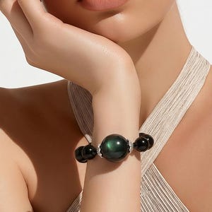 May include: A beaded bracelet featuring a large, central dark green iridescent sphere flanked by smaller black beads and silver-toned metal accents. The bracelet is constructed with a series of smooth, round beads that create a circular band.