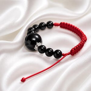 Könnte beinhalten: Ein Armband mit einer Reihe glatter, runder schwarzer Perlen und einer großen zentralen schwarzen Perle, die mit einem silberfarbenen kronenförmigen Zwischenstück verziert ist. Die Perlen sind durch eine rote Kordel mit Schiebeknoten verbunden.
