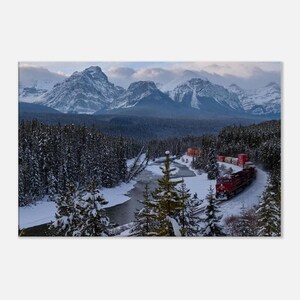 May include: A red freight train travels along tracks winding through a snow-covered evergreen forest. A river flows beside the tracks, and large, jagged, snow-capped mountains rise in the background under a cloudy, overcast sky.