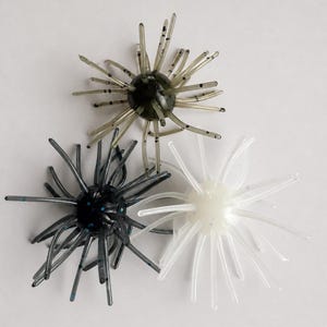 Op de afbeelding: Drie bolvormige viskunstaasjes met uitstralende flexibele tentakels. Eén kunstaas is doorschijnend olijfgroen met donkere spikkels, één is doorschijnend zwart met blauwe spikkels en één is doorschijnend wit met subtiele lichtblauwe spikkels.