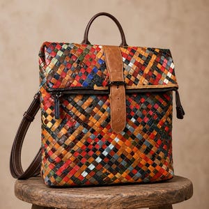 May include: A backpack featuring a multi-colored woven pattern in shades of red, orange, yellow, blue, and green. The bag includes a brown leather top handle, a matching brown leather strap with a buckle closure, a dark zipper, and adjustable straps.