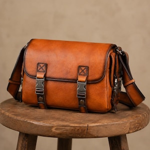 May include: A tan leather messenger bag featuring a rectangular flap, two vertical straps with metallic side-release buckles, and dark brown stitched edges. The bag includes a matching adjustable shoulder strap attached to the sides.