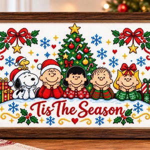 Op de afbeelding: Een ingelijst kruissteekborduurwerk met Snoopy, Woodstock en de Peanuts-bende voor een versierde kerstboom. De tekst Tis The Season is onderaan in het rood geborduurd, omringd door sneeuwvlokken, hulst en cadeautjes.