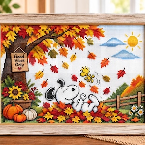 May include: A framed cross-stitch embroidery depicting Snoopy and Woodstock playing in a pile of autumn leaves. The scene features a tree with colorful foliage, a sign reading Good Vibes Only, pumpkins, and a sun in the sky, all encased in a light wood frame.