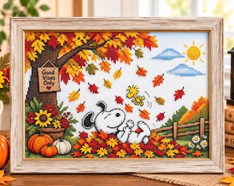 May include: A framed cross-stitch embroidery depicting Snoopy and Woodstock playing in a pile of autumn leaves. The scene features a tree with colourful foliage, a sign reading Good Vibes Only, pumpkins, and a sun in the sky, all encased in a light wood frame.