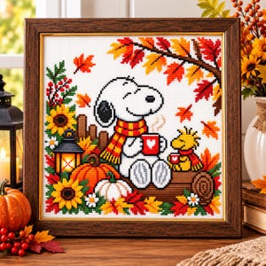 May include: A framed cross-stitch embroidery featuring Snoopy and Woodstock sitting on a log surrounded by autumn leaves, sunflowers, pumpkins, and a lantern. Snoopy wears a red and yellow scarf and holds a red mug with a white heart.