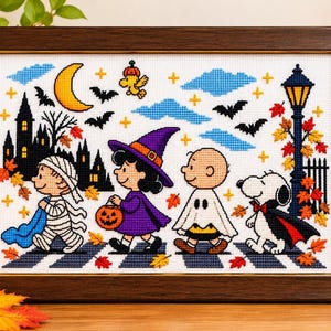 May include: A framed cross-stitch embroidery featuring Peanuts characters in Halloween costumes, including a mummy, a witch, a ghost, and a vampire dog, set against a night scene with a crescent moon, bats, and a street lamp.