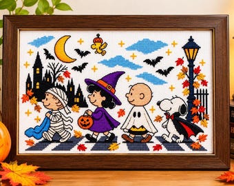 May include: A framed cross-stitch embroidery featuring Peanuts characters in Halloween costumes, including a mummy, a witch, a ghost, and a vampire dog, set against a night scene with a crescent moon, bats, and a street lamp.