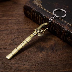 Puede incluir: Un llavero de metal color latón con forma de empuñadura de espada y vaina. La vaina presenta el texto "進撃の巨人" y "Attack on Titan" impreso en negro. El llavero incluye una anilla metálica circular unida por una cadena corta.