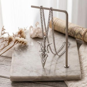 May include: A silver-toned metal pendant shaped like a stylised spearhead or trident, suspended from a matching silver-toned link chain. The pendant features a pointed tip, a central shaft, and curved side elements, all crafted with a polished finish.