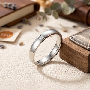 Op de afbeelding: Een gepolijste zilverkleurige metalen ring met een enkele, kleine, ronde, heldere gefacetteerde edelsteen die verzonken in het midden van de band is geplaatst. De ring heeft een glad, reflecterend oppervlak en een eenvoudig, doorlopend rond ontwerp.