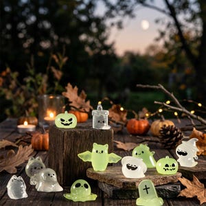 Könnte beinhalten: Eine Sammlung kleiner, im Dunkeln leuchtender Halloween-Figuren, darunter Geister, eine Fledermaus, ein Kürbis, eine Kerze und ein Grabstein. Die durchscheinenden, blassgrünen Figuren haben einfache schwarze Gesichter und stehen auf Holz.